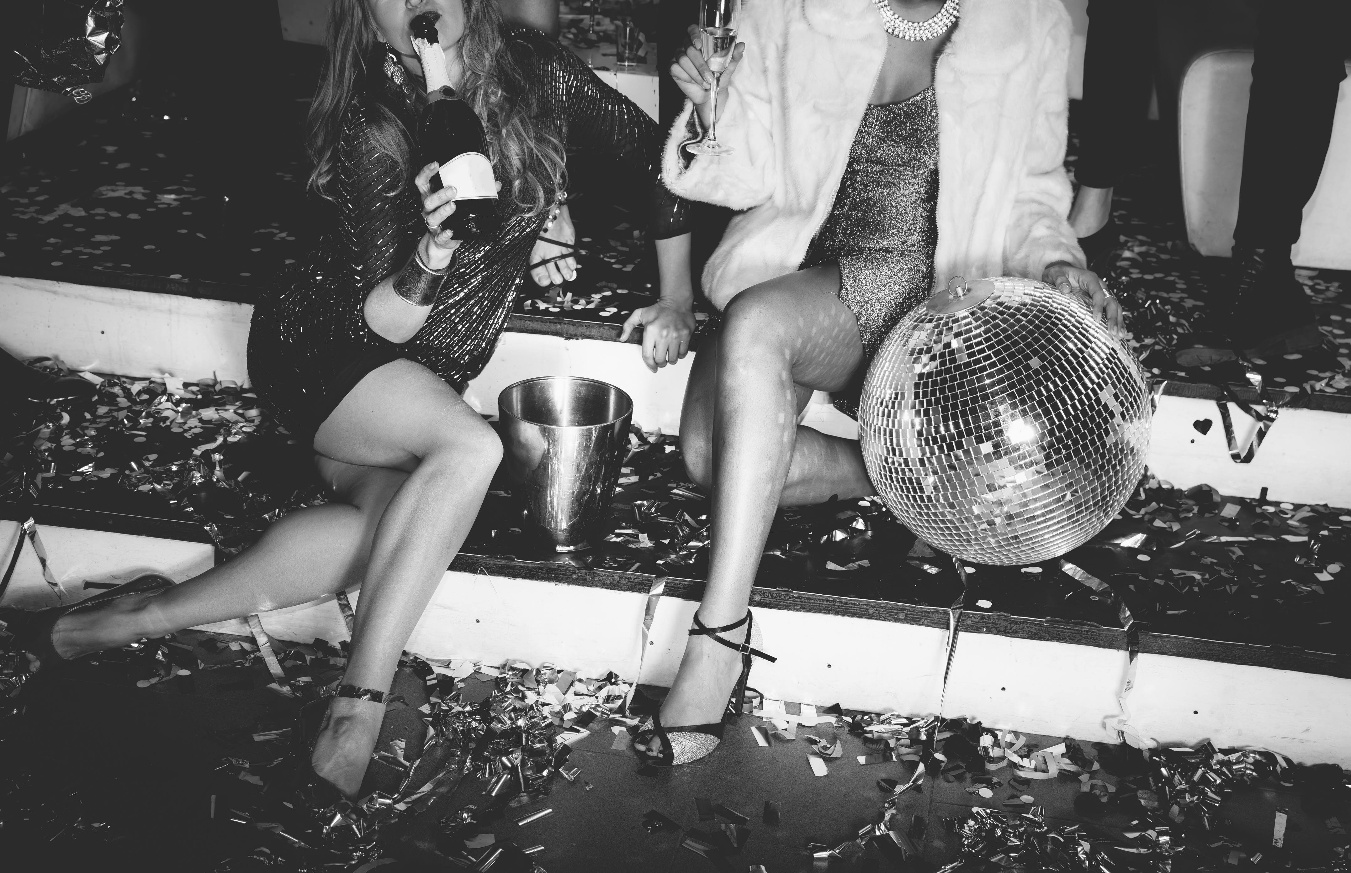 Two women sitting down with a bottle of champagne as a party rages around them.