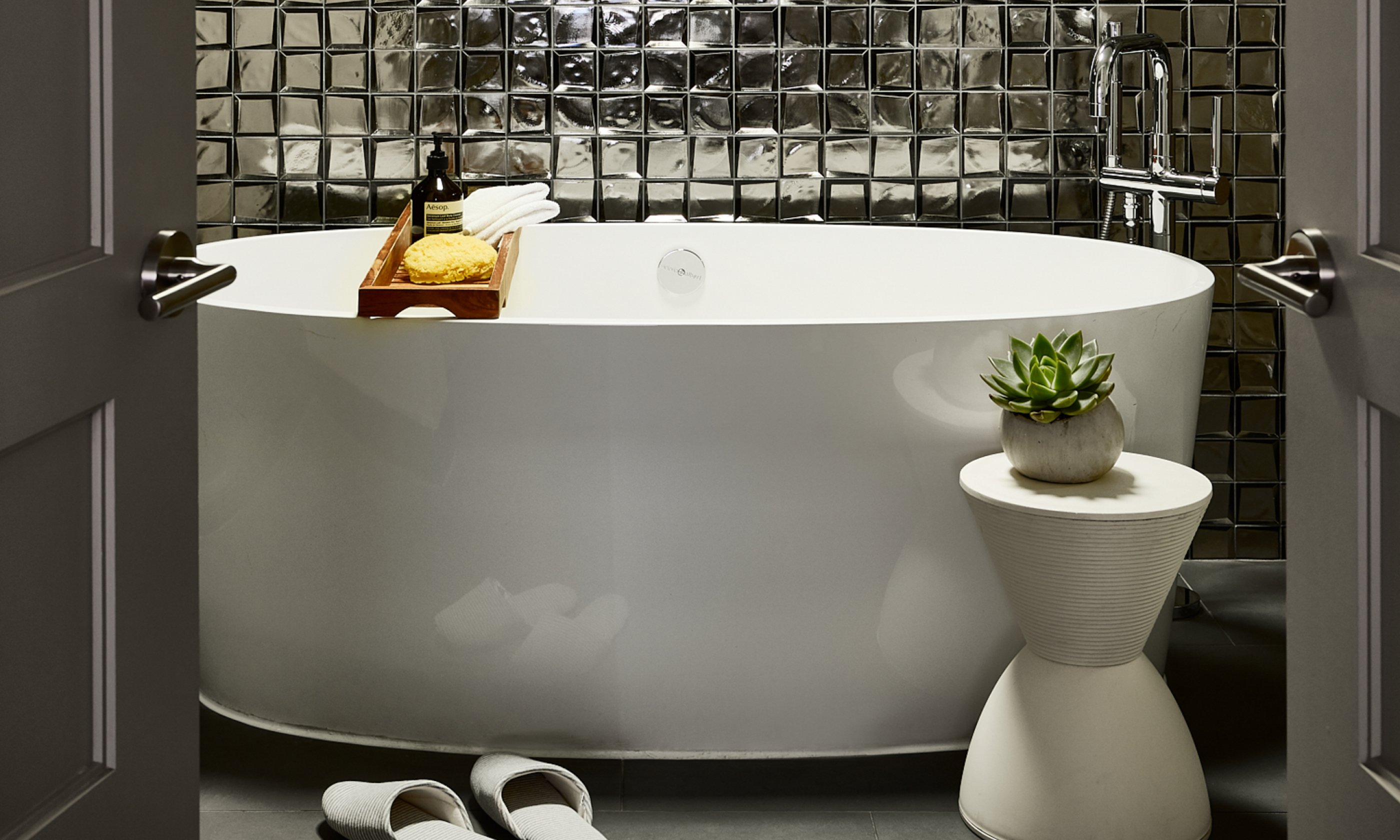 Metallic tiled bathroom with oversized soaking tub and white flip flops.