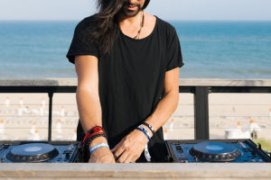 A man in a black tee shirt DJs at a turntable with a view of the ocean in the background.