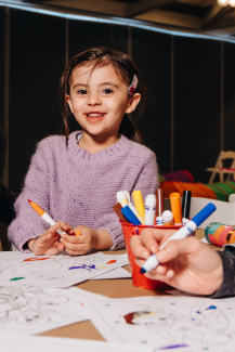 A young girl smiles at the camera while coloring a worksheet