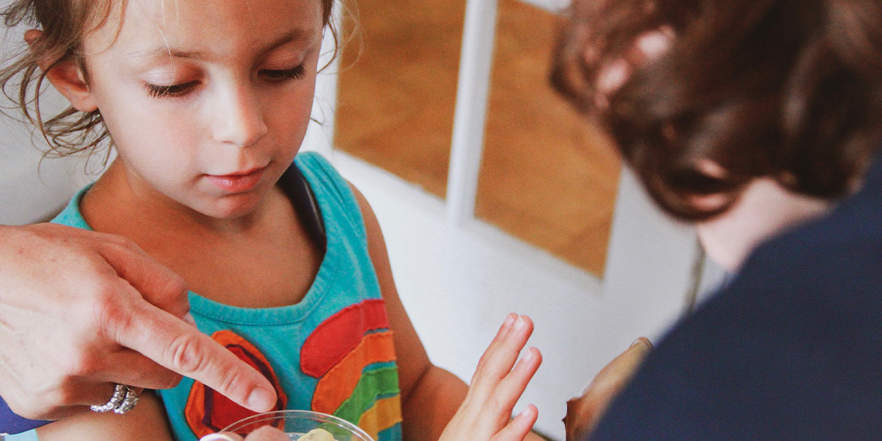 Child in a colorful shirt holding up hands, with an adult pointing at their fingers, in a bright room.