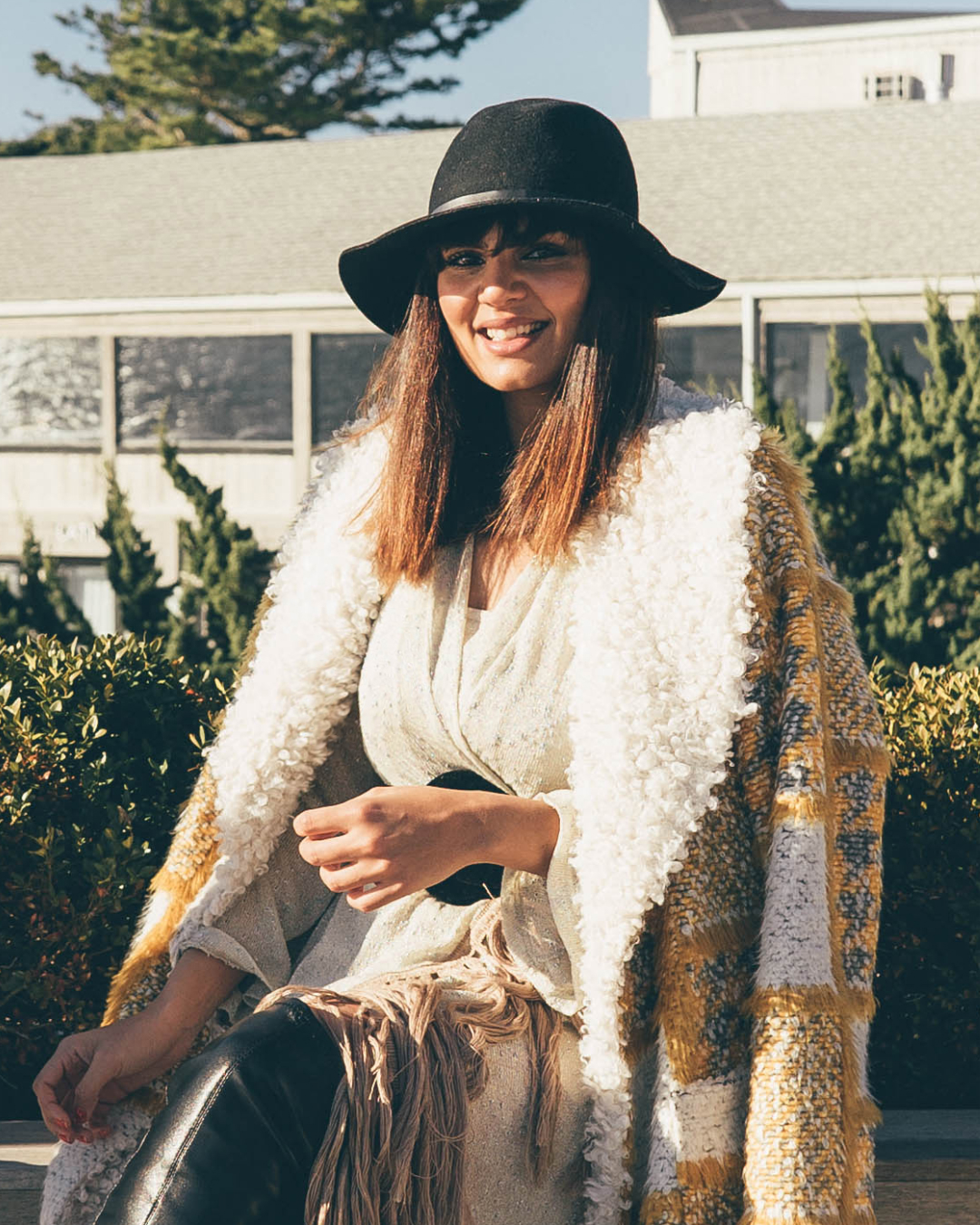 Woman in a white sweater and black hat smiles outdoors, with greenery and houses in the background on a sunny day.