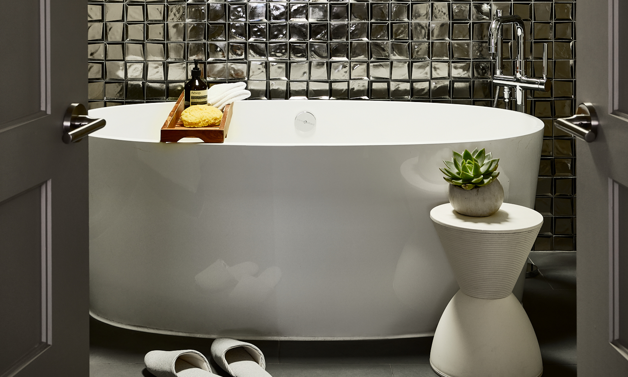 Metallic tiled bathroom with oversized soaking tub and white flip flops.