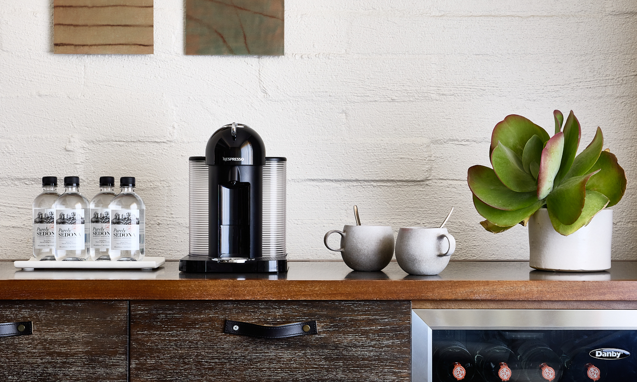 Mini fridge and counter with waters, Nespresso machine and two coffee cups.