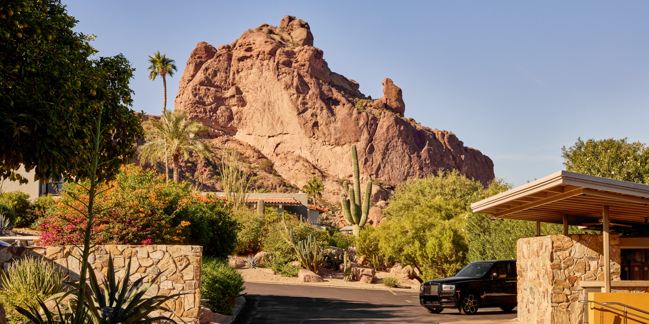 Desert landscape with a prominent rocky mountain in the background, surrounded by lush greenery and a parked black car in the foreground.