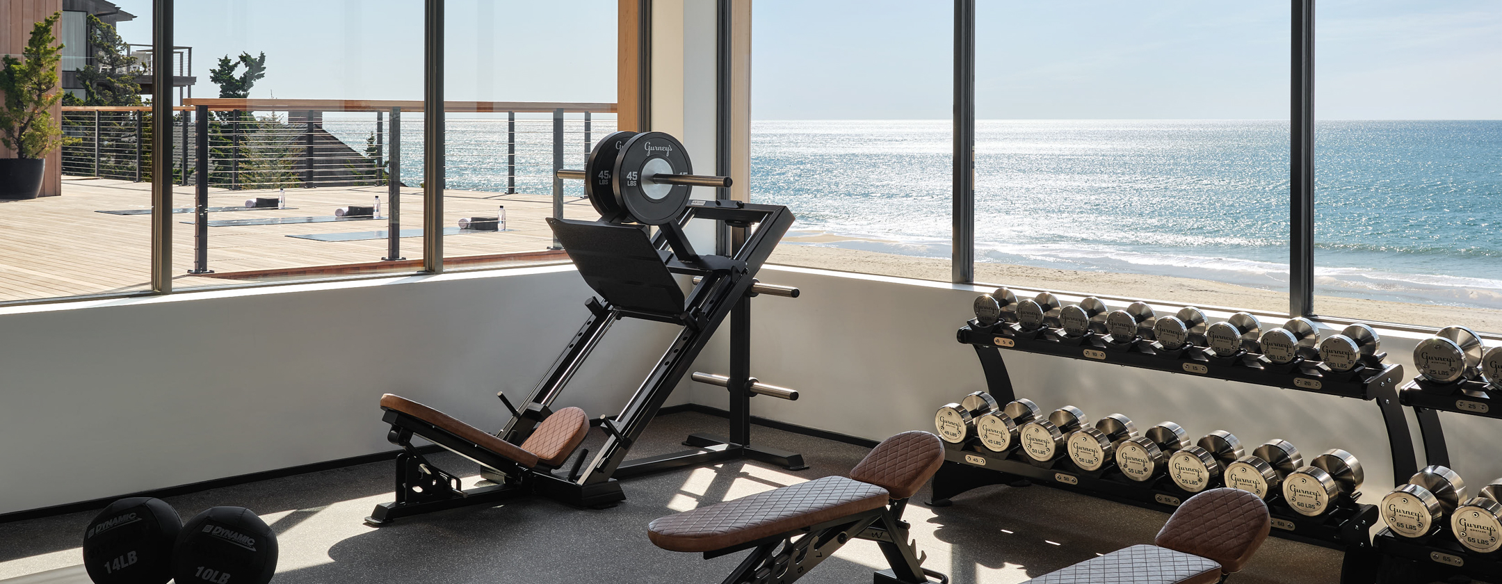 A gym with exercise equipment and a large window showing a beach view.