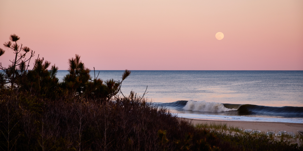 View from the beach of the moon on the horizon over the ocean at dusk.