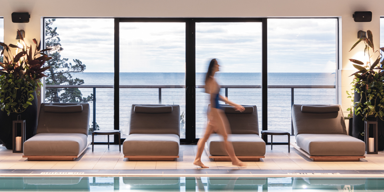 Woman walking by indoor pool with lounge chairs and beach view in background.