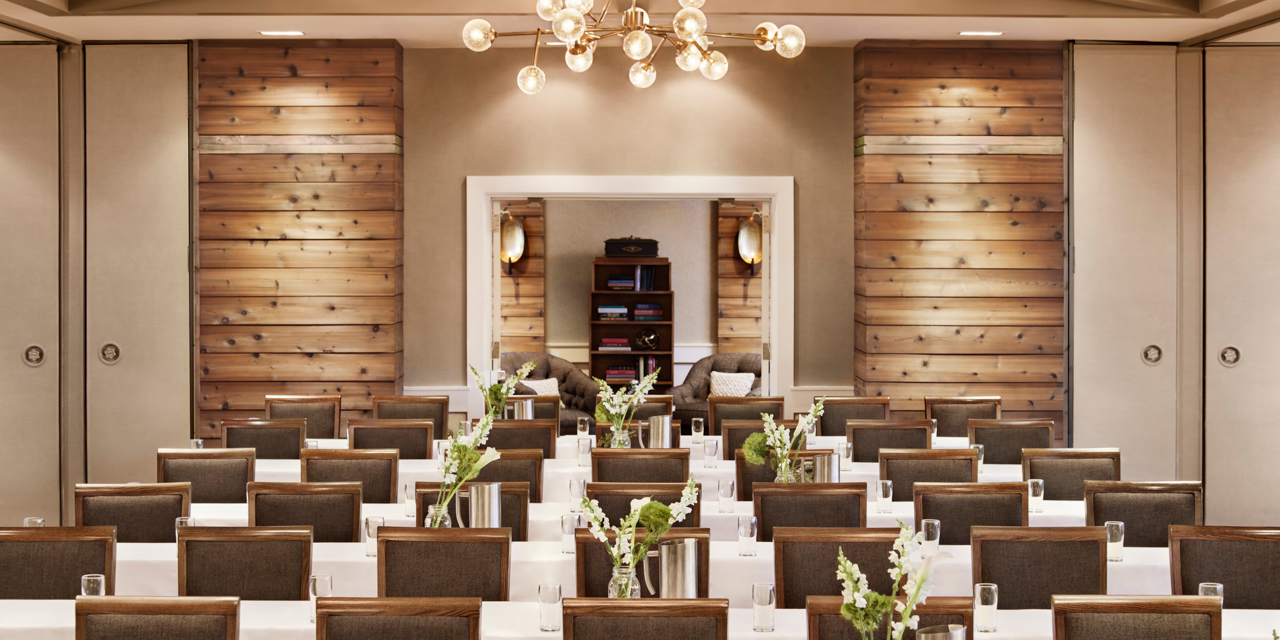 Meeting space set up with podium and rows of tables and chairs with wood wall accents and chandelier.