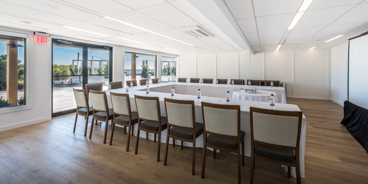 Latitude with wood floor, white walls, u-shaped table setup with white linens, projector and screen.