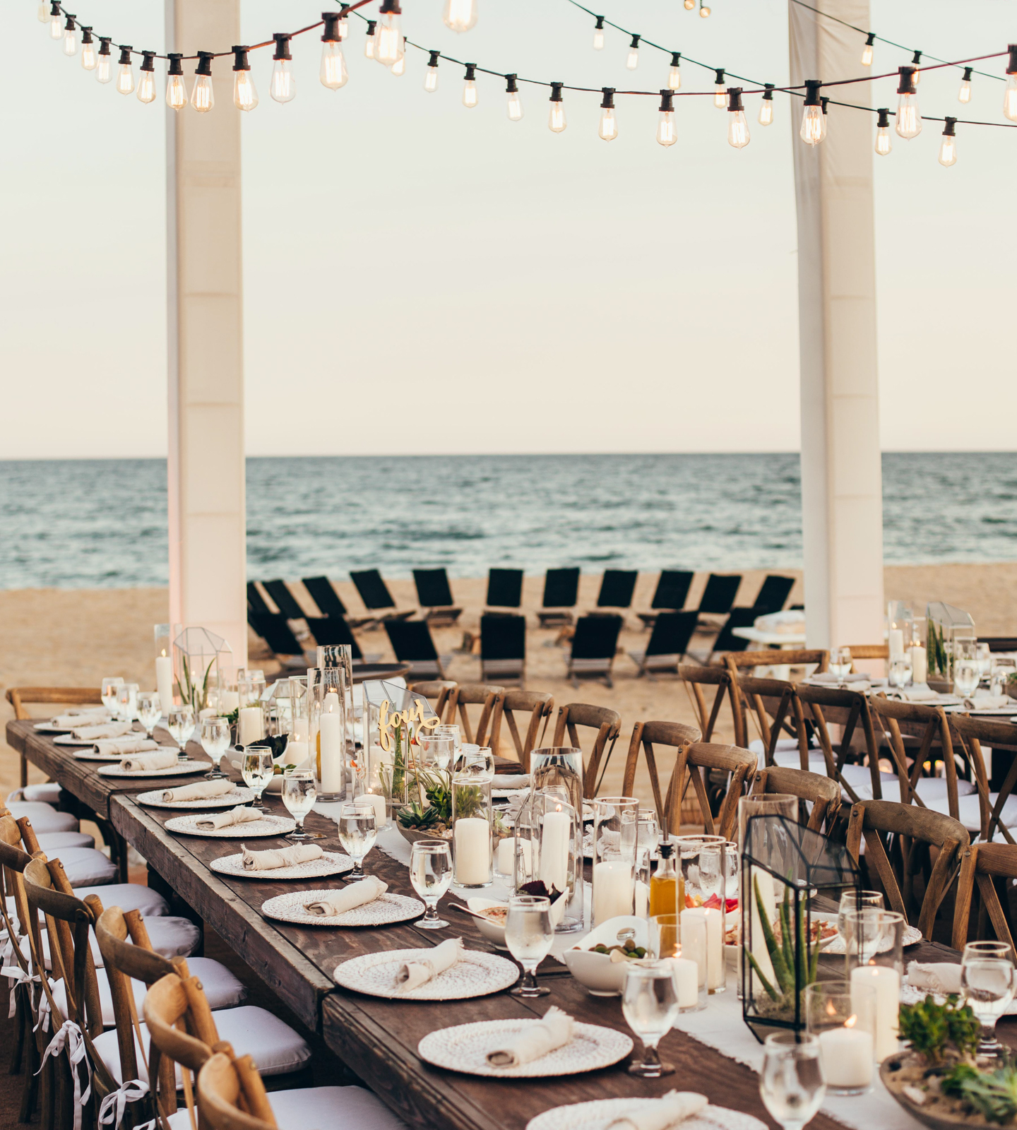  View from beach-side long wooden table set for dinner with hanging lights, candles and plant center pieces as sun sets with beach fire-pit in background.