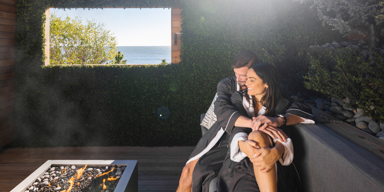 Couple in robes cuddling by outdoor fire pit at gurney's seawater spa.