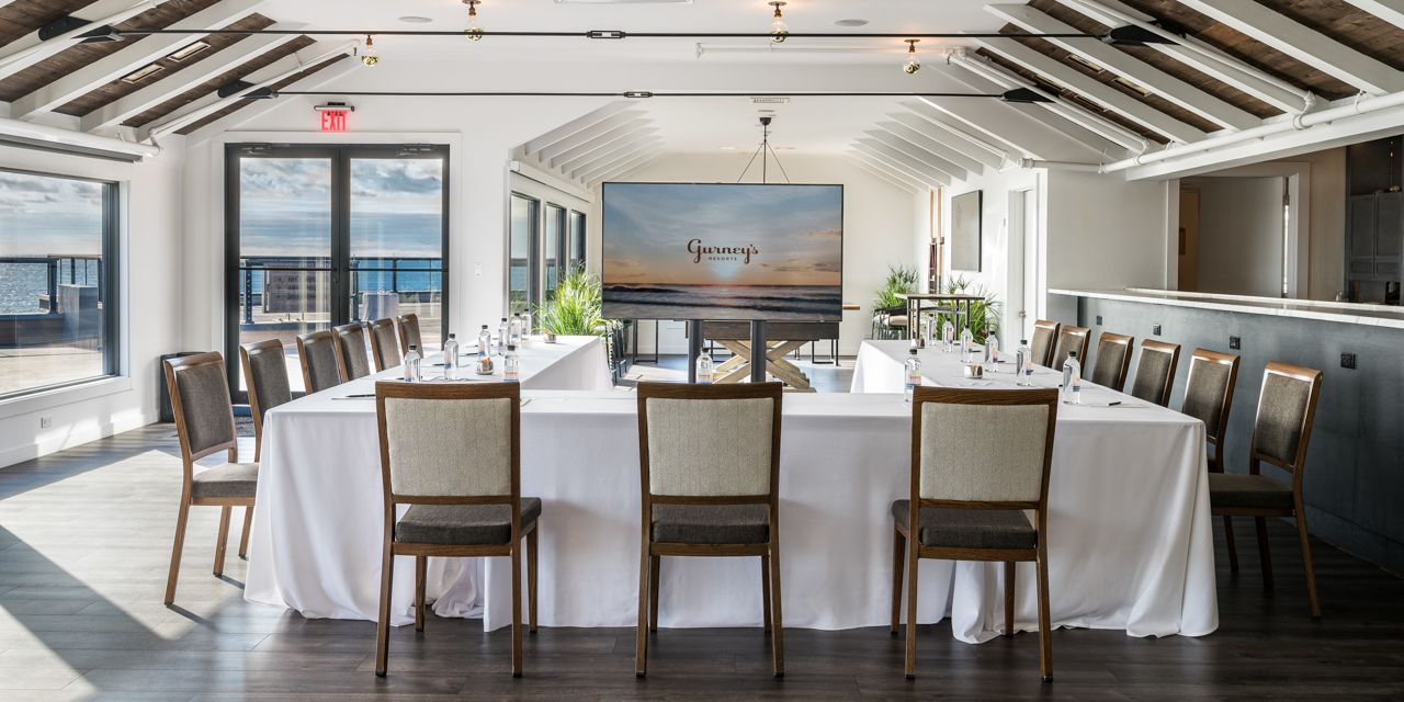 The Regent Lounge with wood floors, white walls and ceiling with wooden accents, u-shaped table setup with white linen and television with Gurney’s Logo on screen.