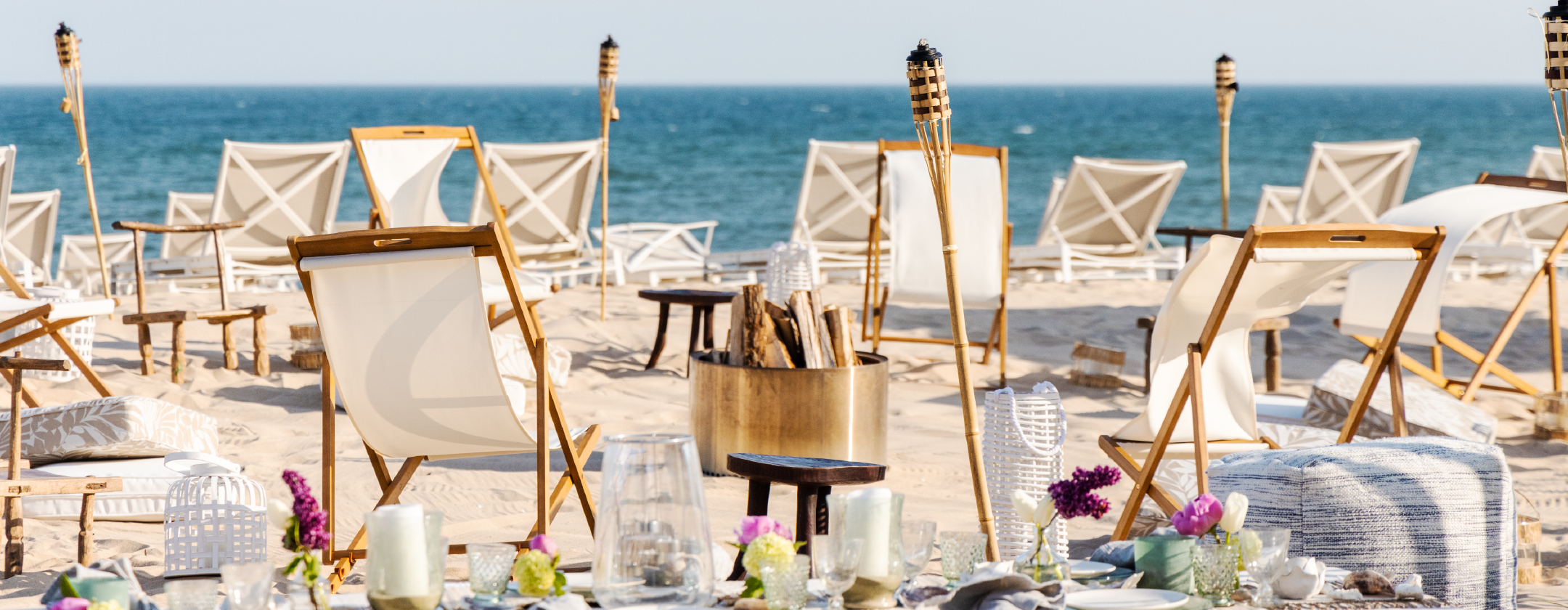 A beach setup with white chairs, a table set with glasses and flowers, and a view of the ocean.