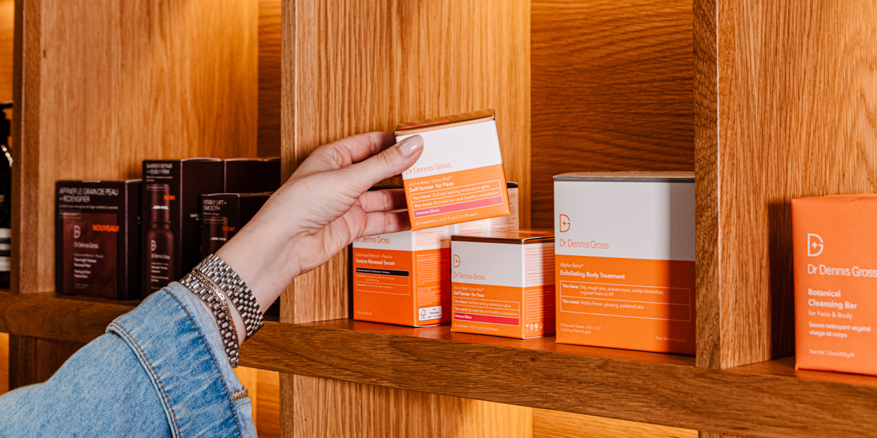 Woman in denim long sleeve picking up Dr. Dennis Gross product from wood shelf in orange and white packaging.