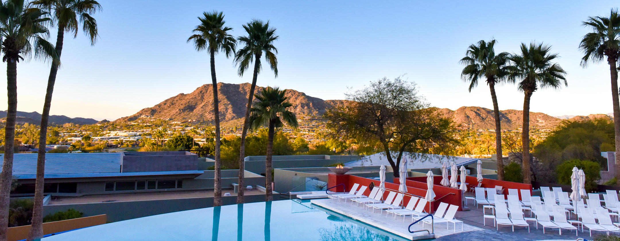 View of Infinity Pool with white lounge chairs and umbrellas as sun sets over mountain in background.