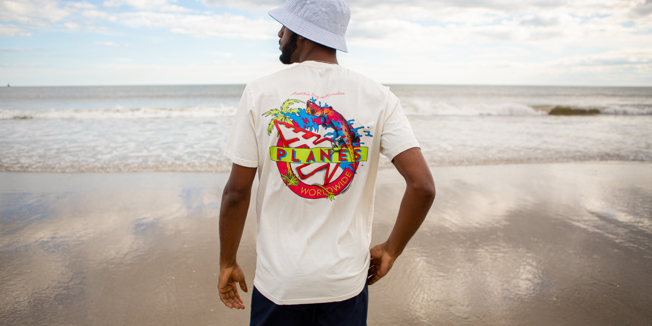 A man stands with his back toward the beach in a white t-shirt and bucket hat from Paper Planes.
