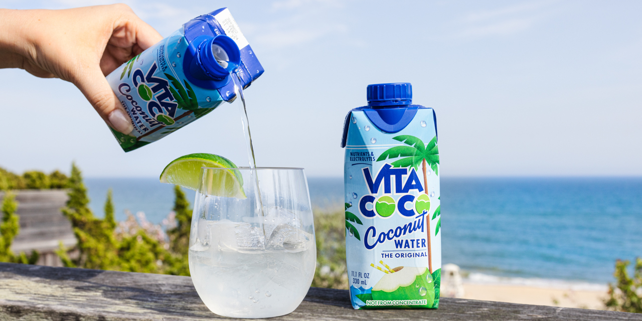 Coco Vita being poured into cocktail glass with ice and lime wedge with beach in backdrop.