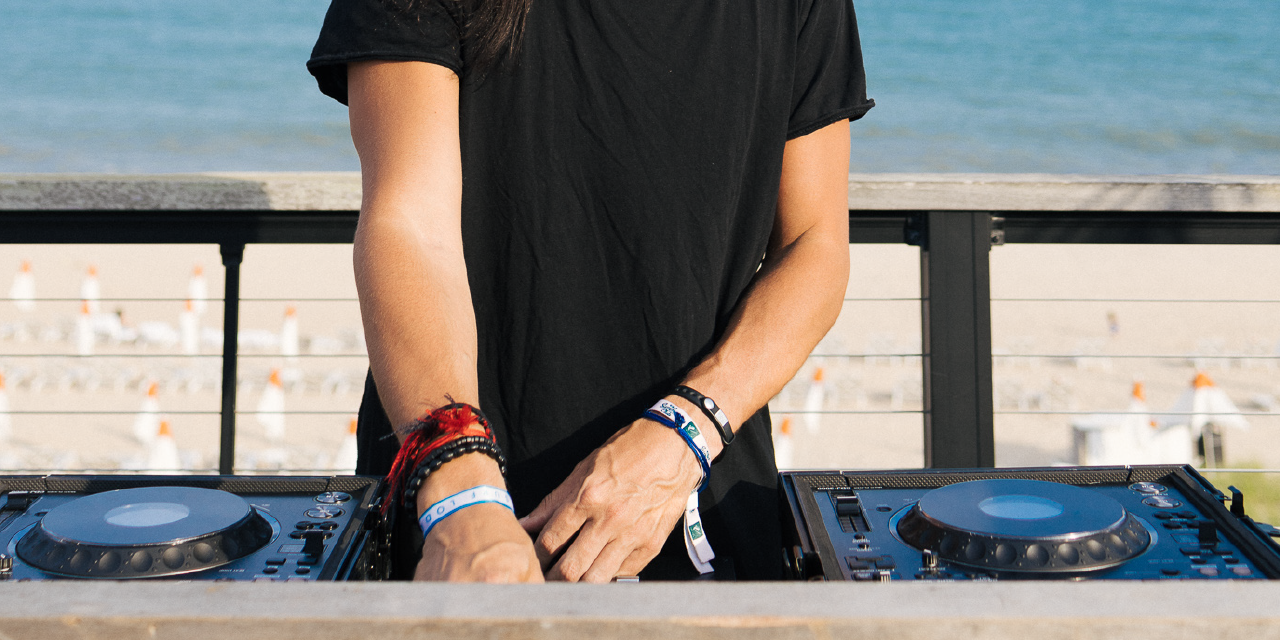 A man in a black tee shirt DJs at a turntable with a view of the ocean in the background.