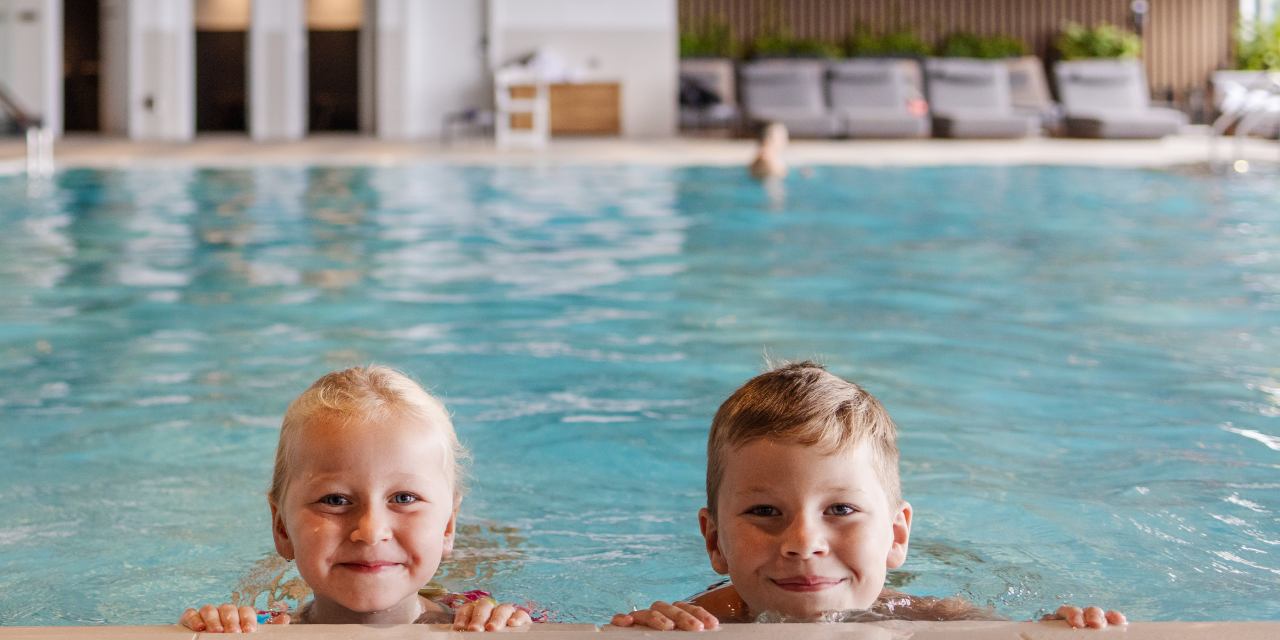 Two children smile at the edge of an indoor pool