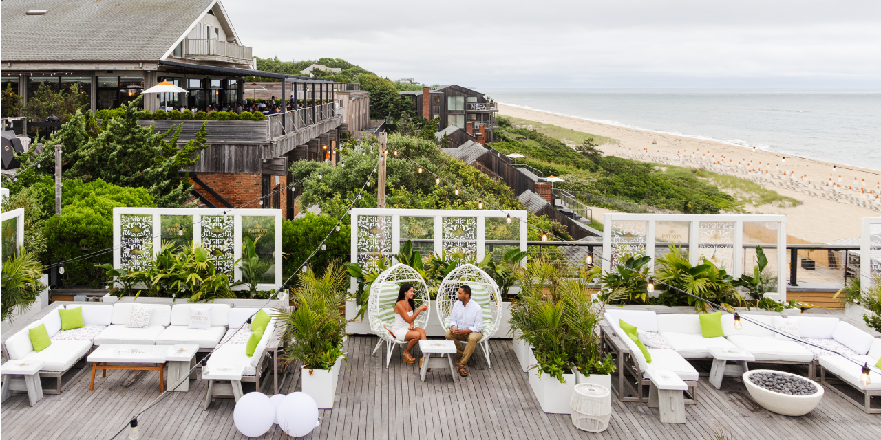 A couple sits drinking cocktails in a plant-filled space with the ocean in the background