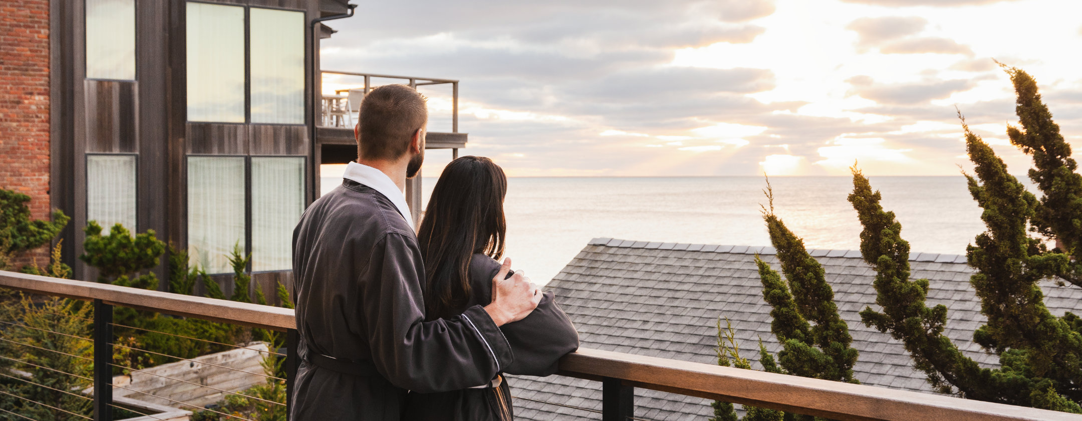 A couple in robes stands on a balcony overlooking the ocean at sunset.