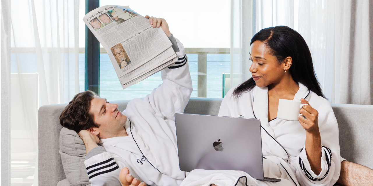 A couple in robes read the newspaper and browse on a laptop with the ocean in the background