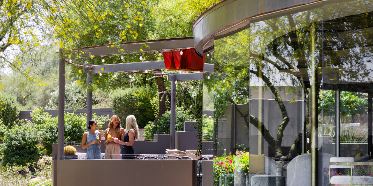 Three girlfriends enjoying craft cocktails and a laugh on jade bar patio surrounded by lush desert landscape.