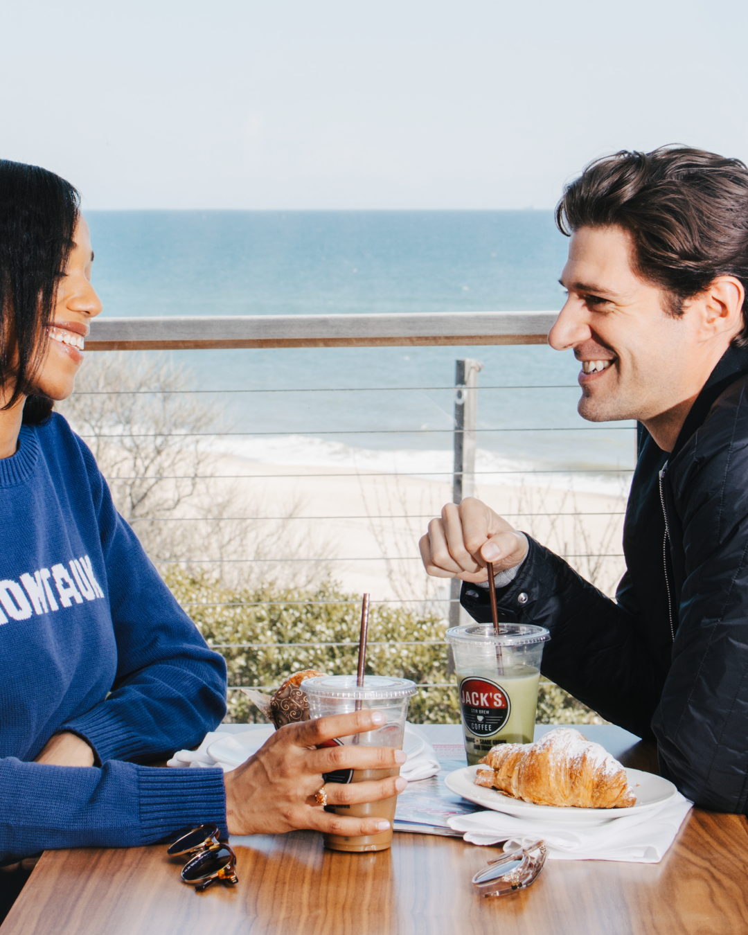 A man and woman hold colorful drinks in front of an ocean backdrop