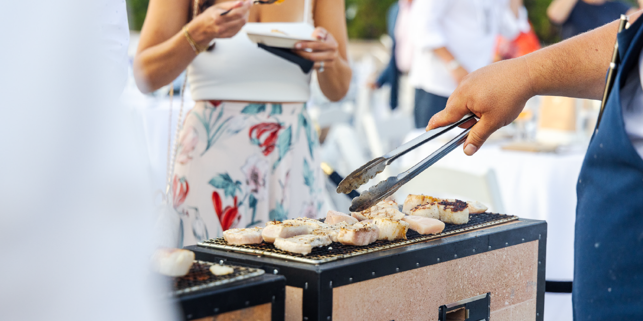 A woman stands in front of a grill as a hand turns clams with tongs