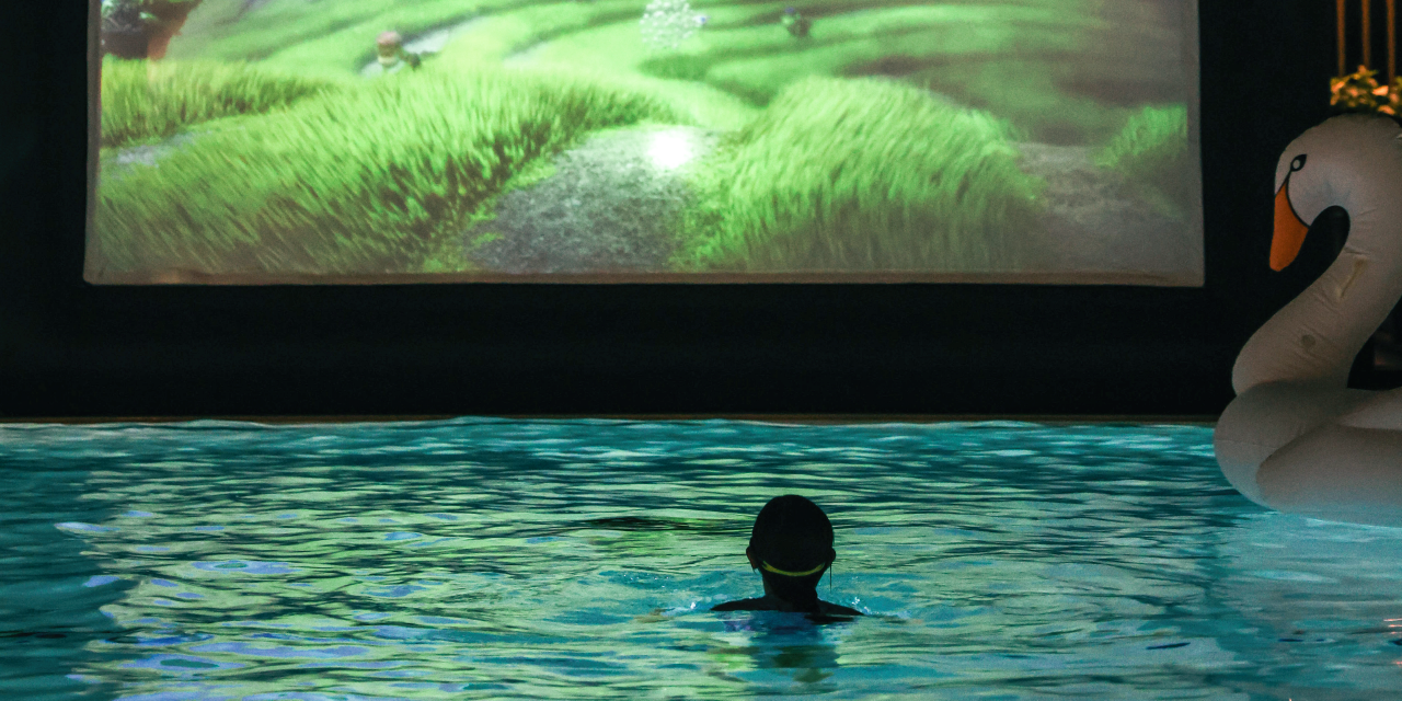 A child bobs in a pool in front of a bright screen