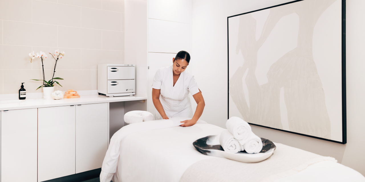 A woman in scrubs flattens a blanket in a white spa treatment room