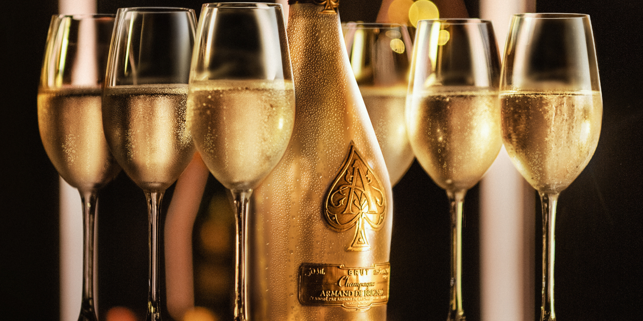 Gold bottle of Armand de Brignac Champagne against black background surrounded by filled glasses of champagne.