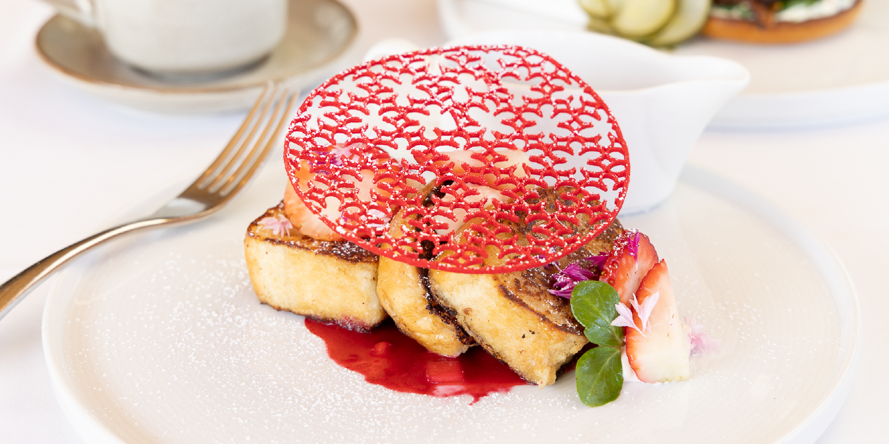 Strawberry waffles with a red lace garnish and strawberry compote on the side against a white tablecloth