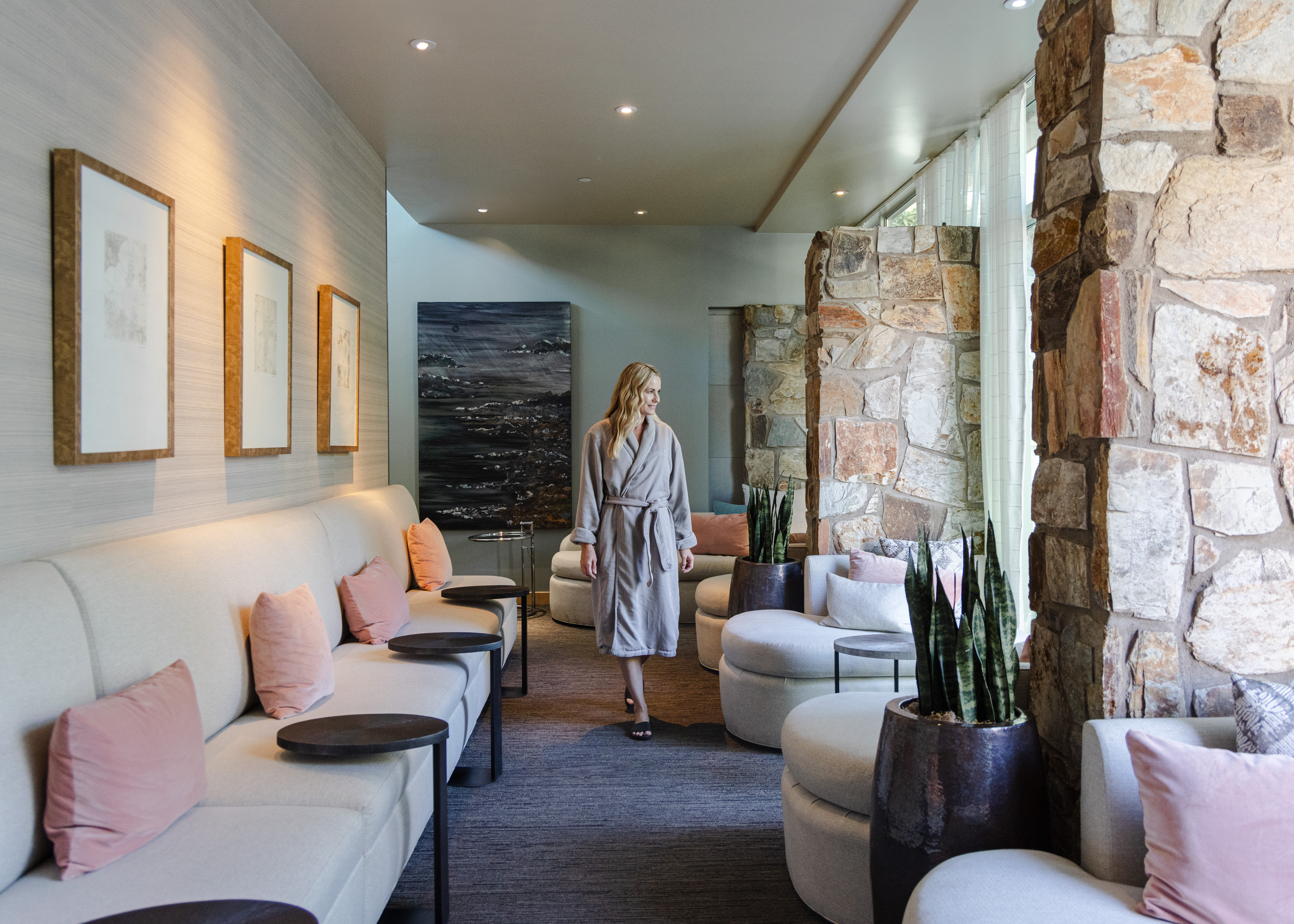 Woman in robe walking through a modern spa lounge with beige sofas, pink cushions, and stone walls.