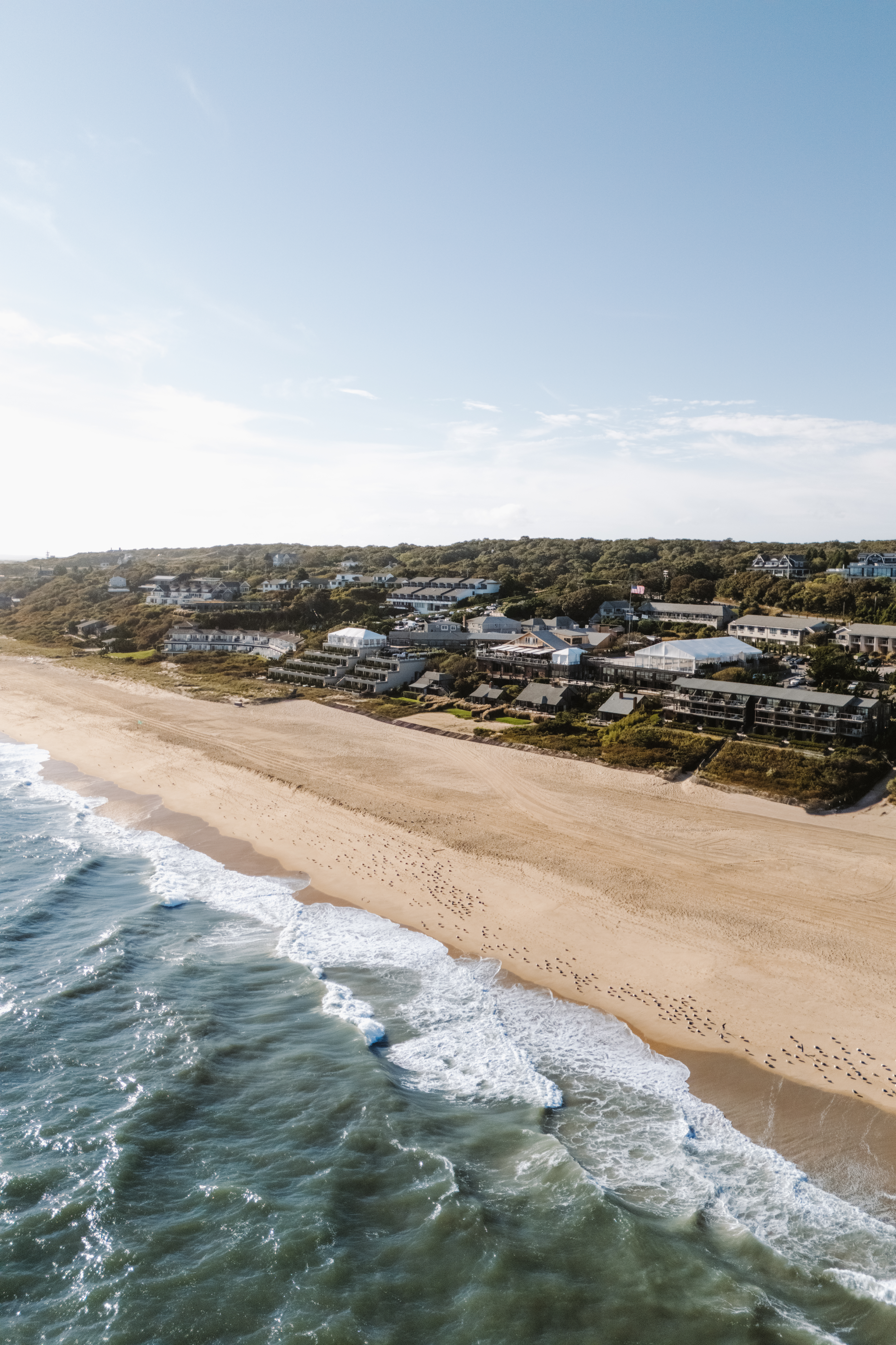 Coastal aerial view with waves crashing on a sandy beach, adjacent to a town with houses and greenery under a clear blue sky.
