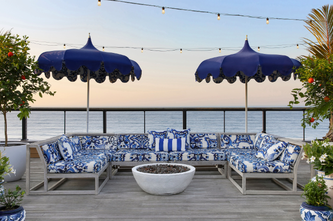 Cozy outdoor lounge with blue-patterned seating, two navy umbrellas, overlooking the ocean at sunset. String lights hang above.