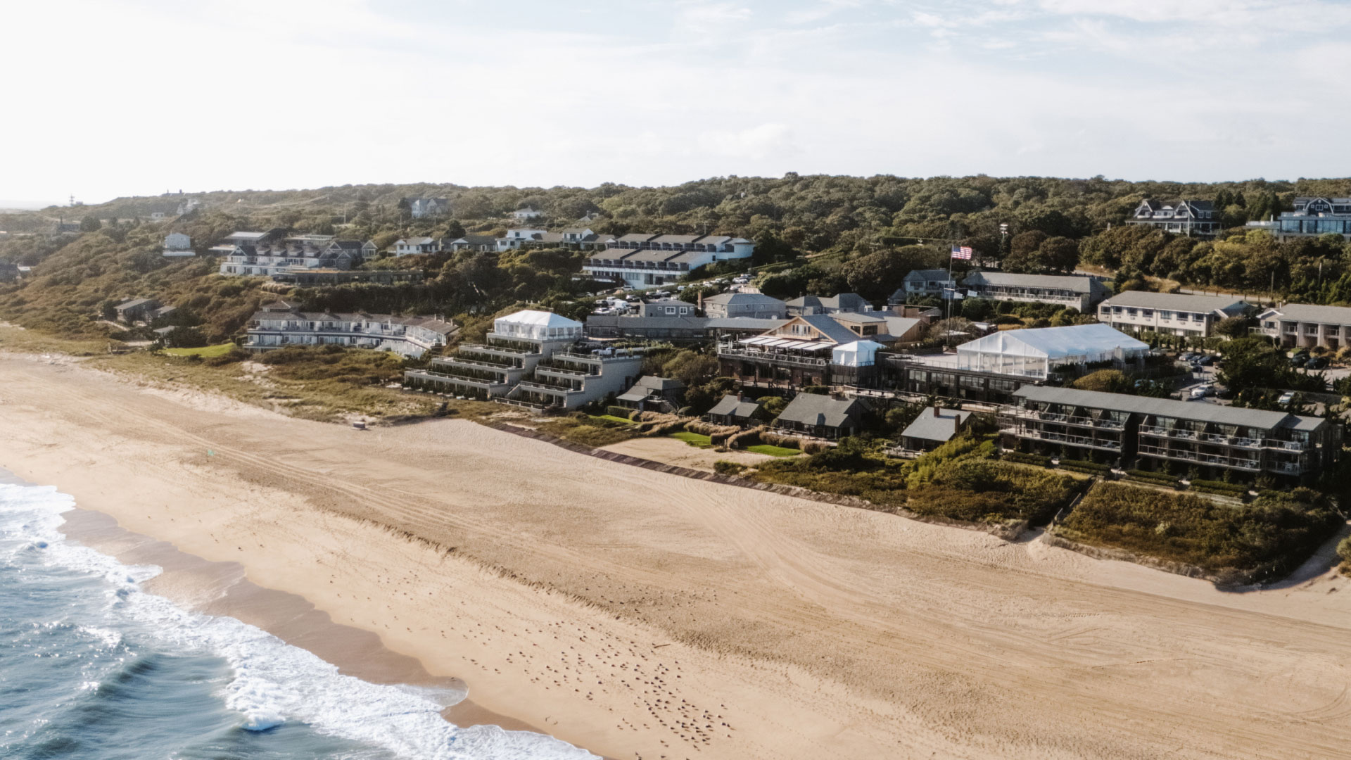 Aerial view of a sandy beach with waves, backed by a coastal town with green trees and buildings in the background.
