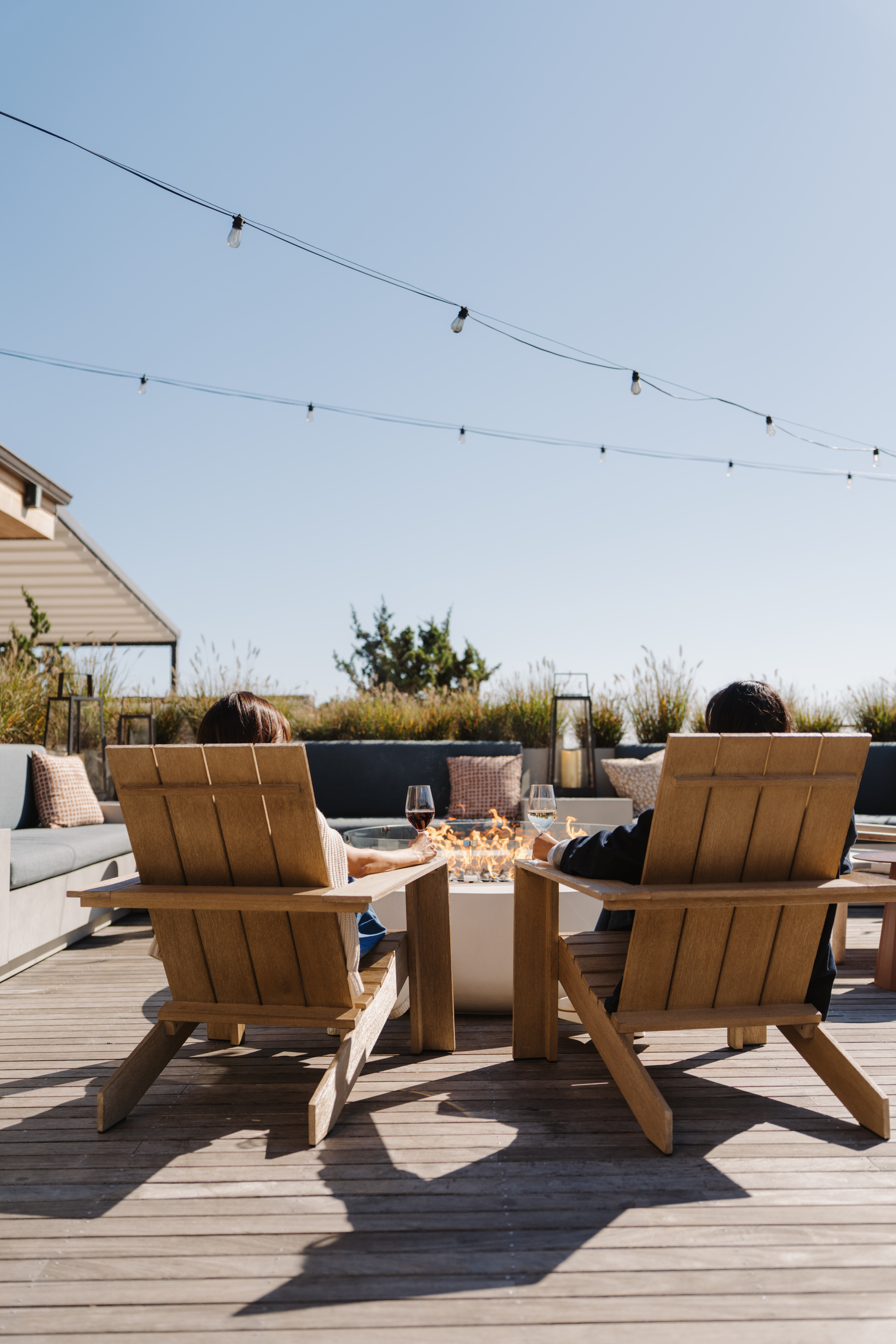 Two people relaxing on wooden chairs in a sunny outdoor setting, with string lights overhead and blue sky.