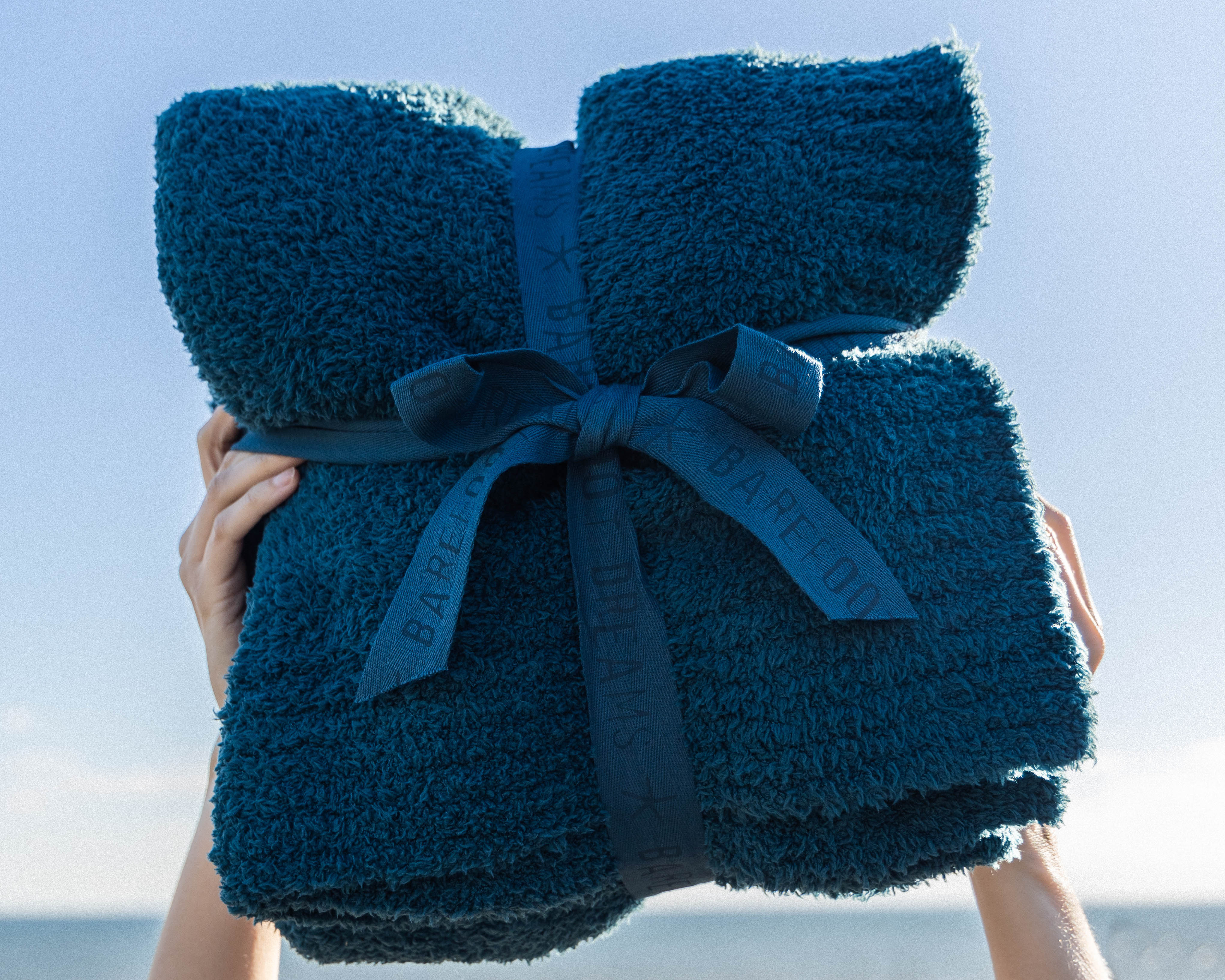 Woman holding blue plush Barefoot Dreams blanket on the beach.