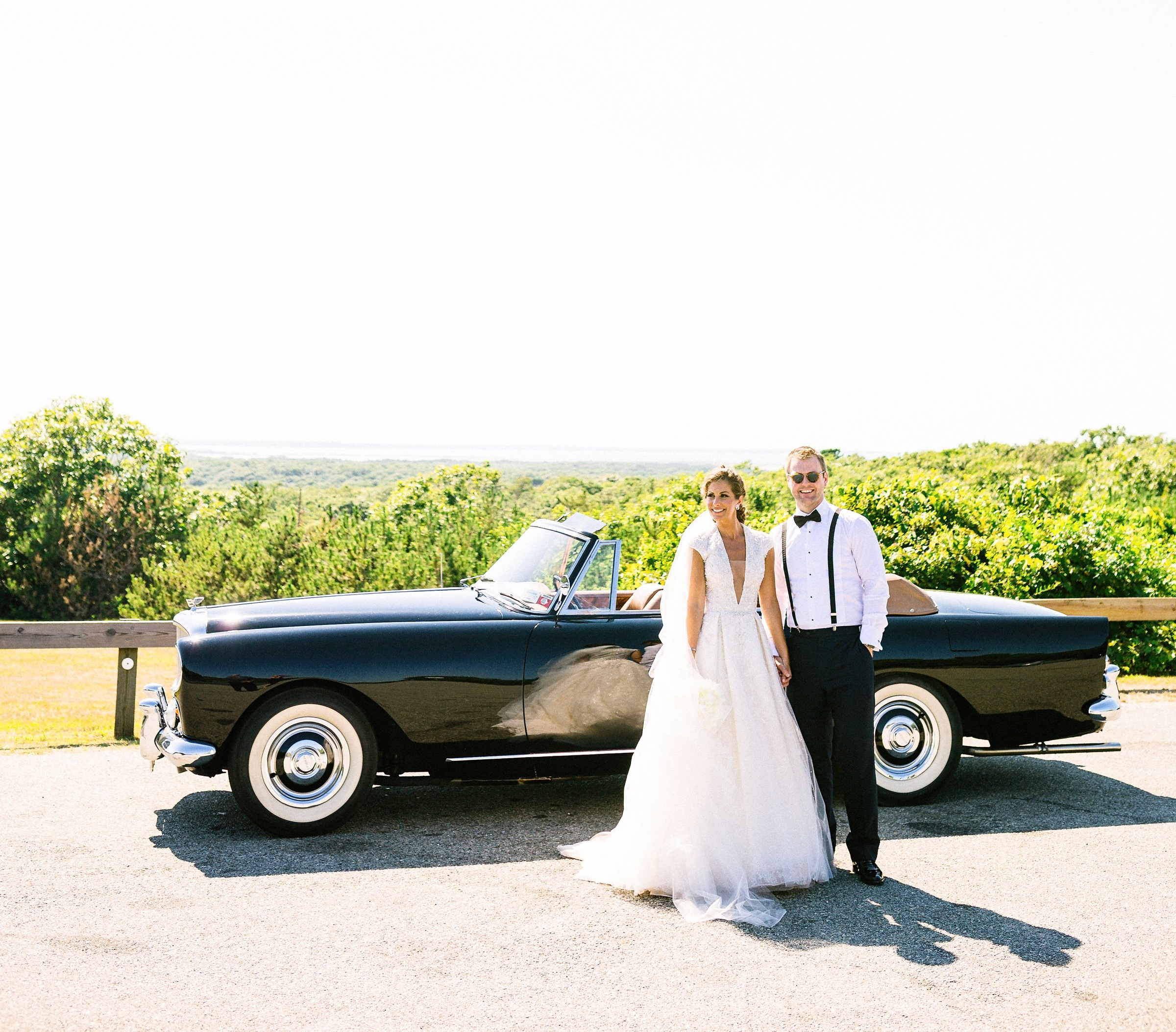 Bride and groom standing in front of black convertible with beach and shoreline in background.