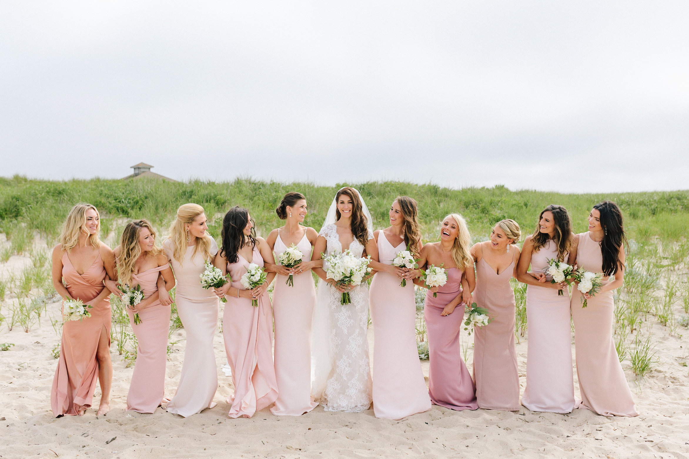 Bride and her bridesmaids in light pink dresses standing in a line on the beach.