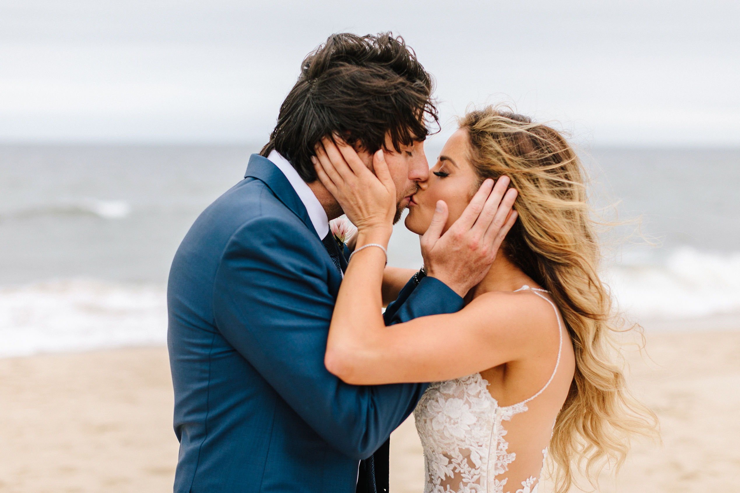 Bride and groom kissing on the beach with the shoreline in background.