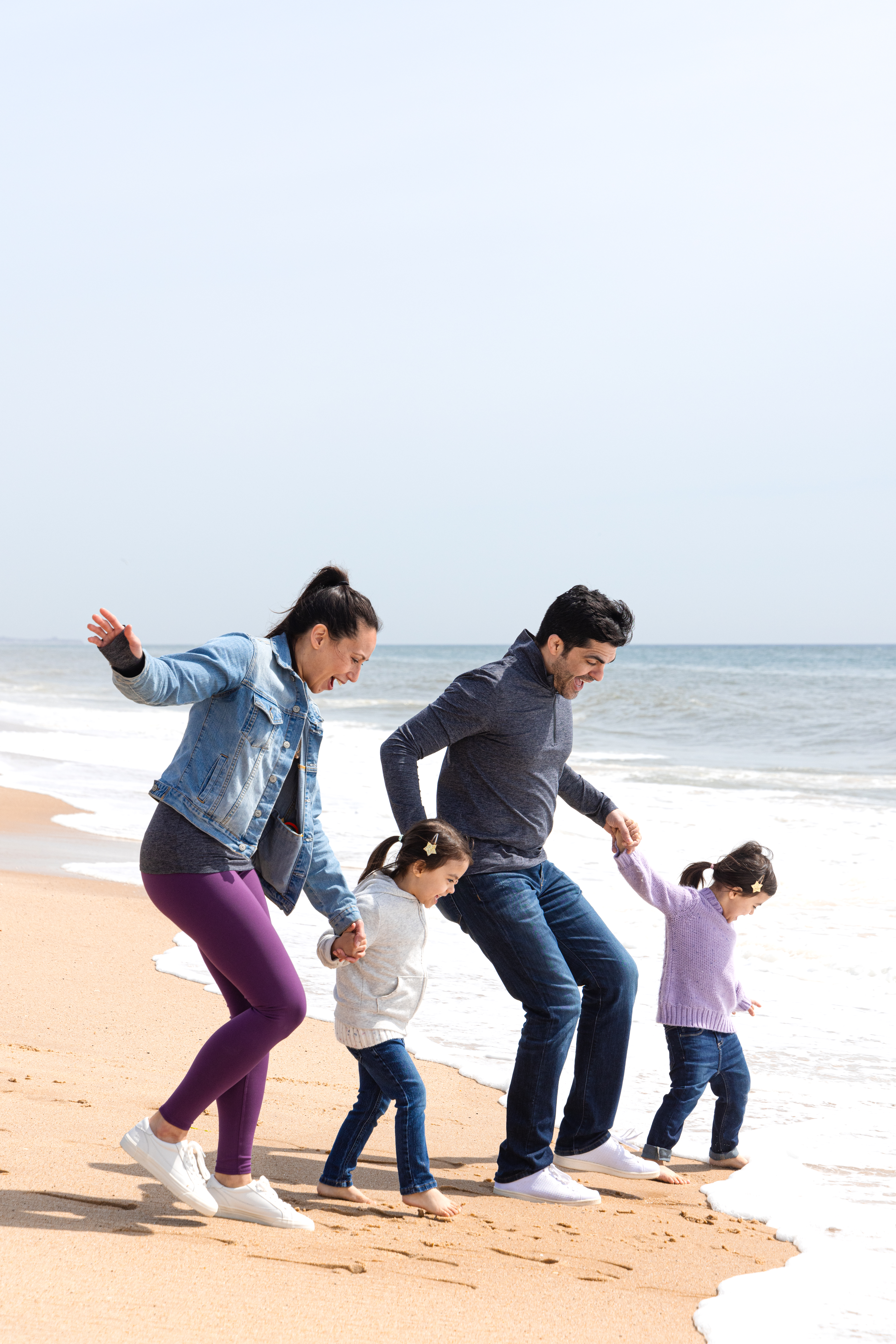 Husband, wife and two daughters laughing as they run through the water on the beach.