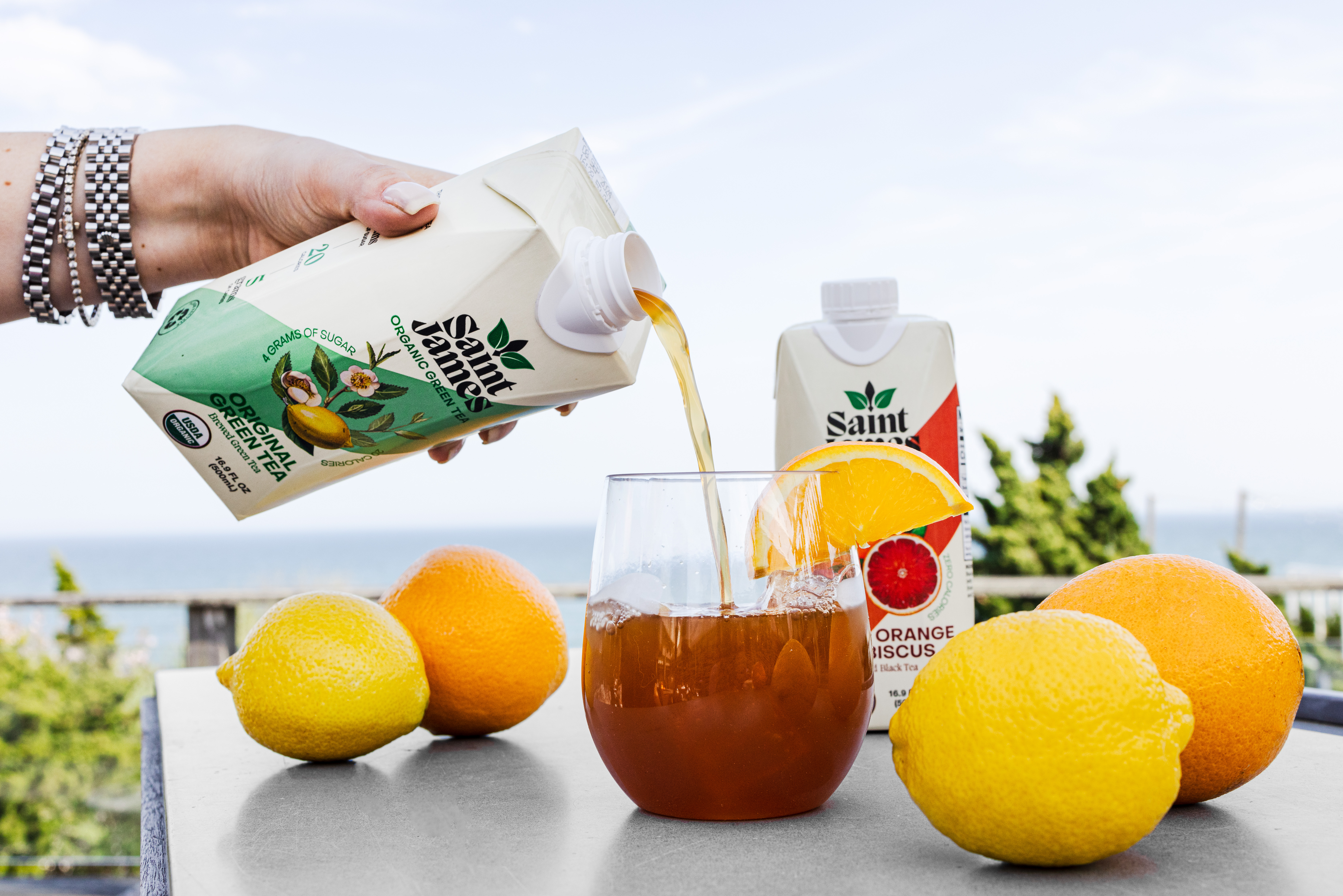 Saint James Iced Tea being poured into a glass surrounded by citrus with beach in background.