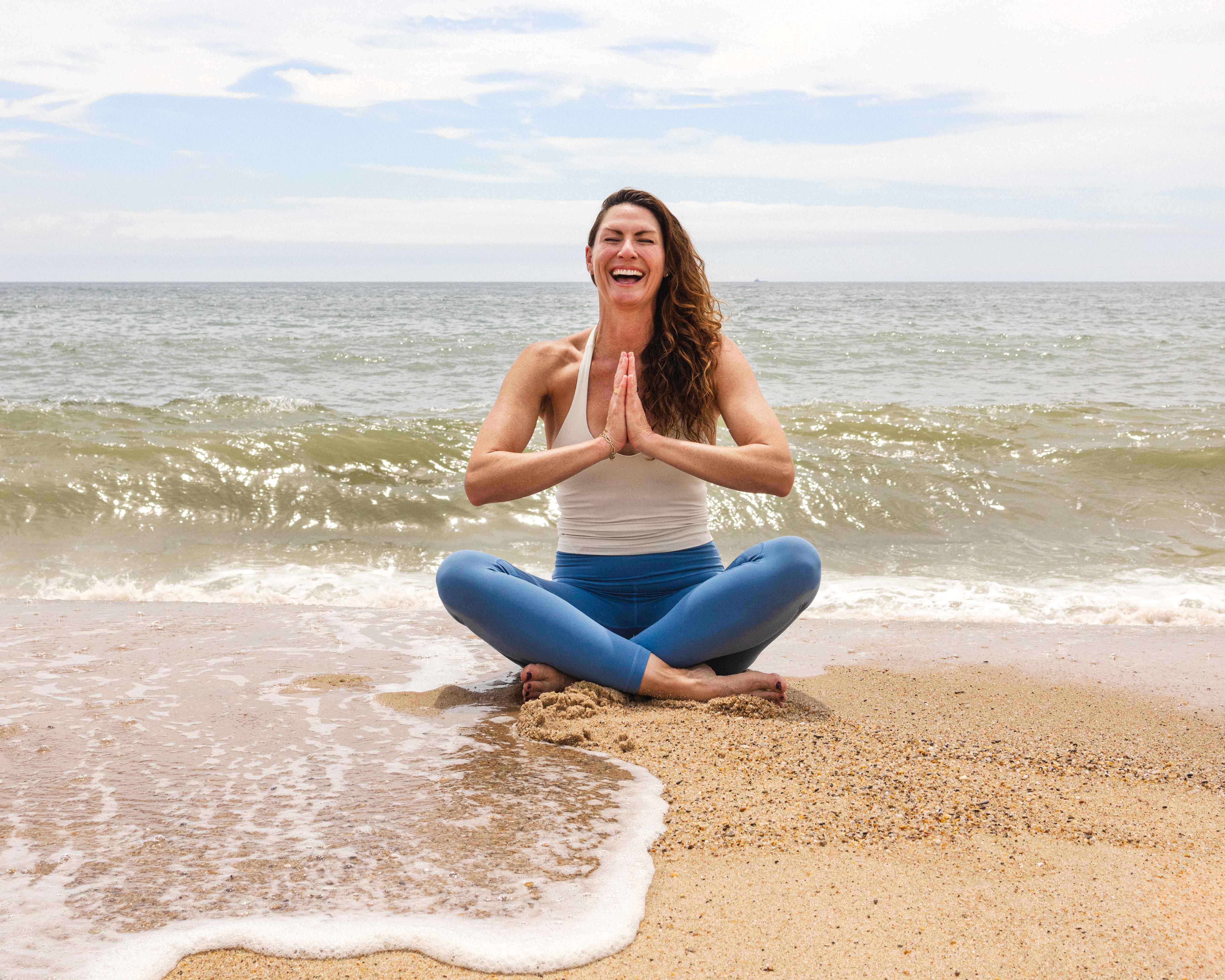 Kat Ruiz smiling and sitting in yoga pose on beach as waves break around her.