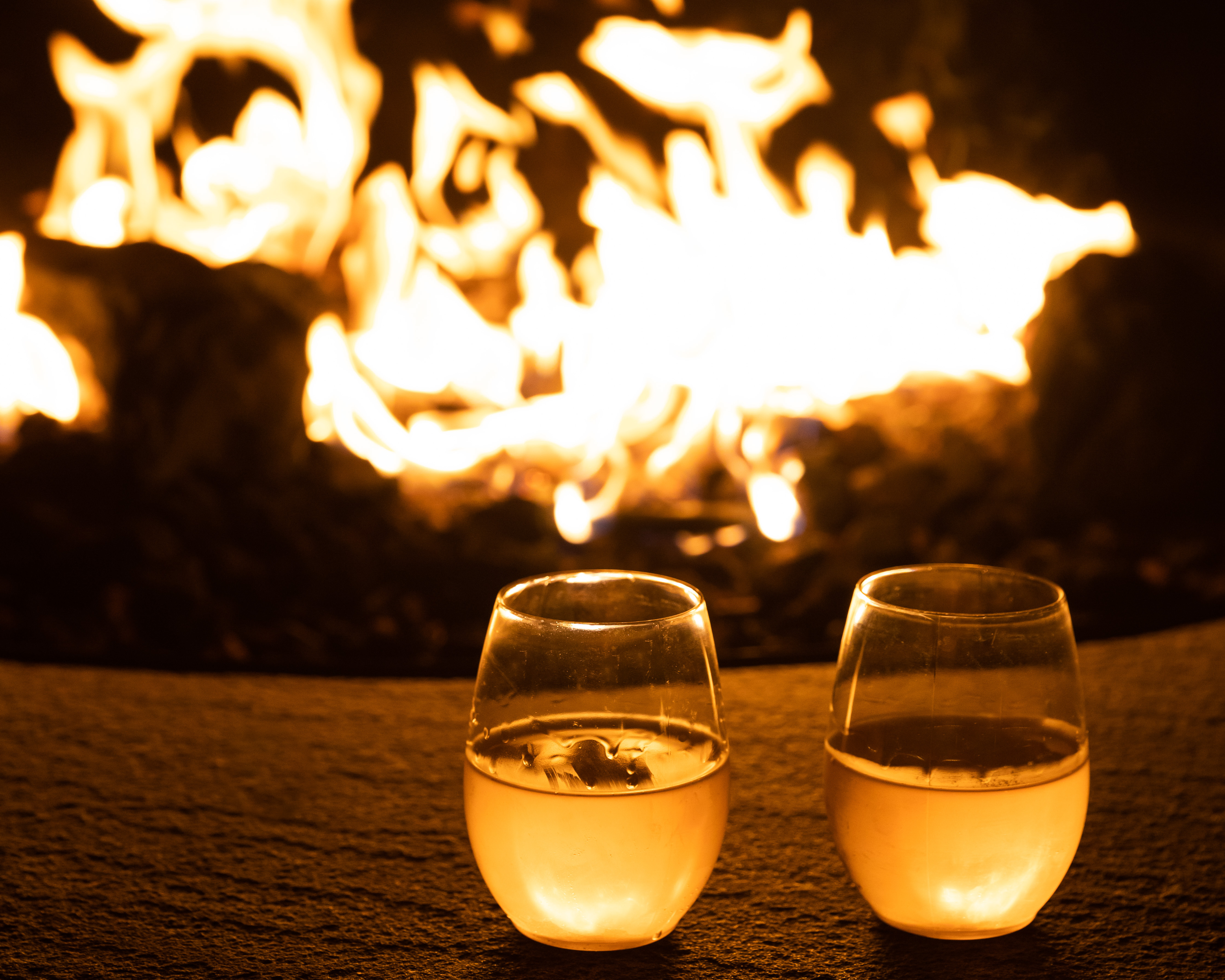 Two glasses of wine in front of fire pit at night.