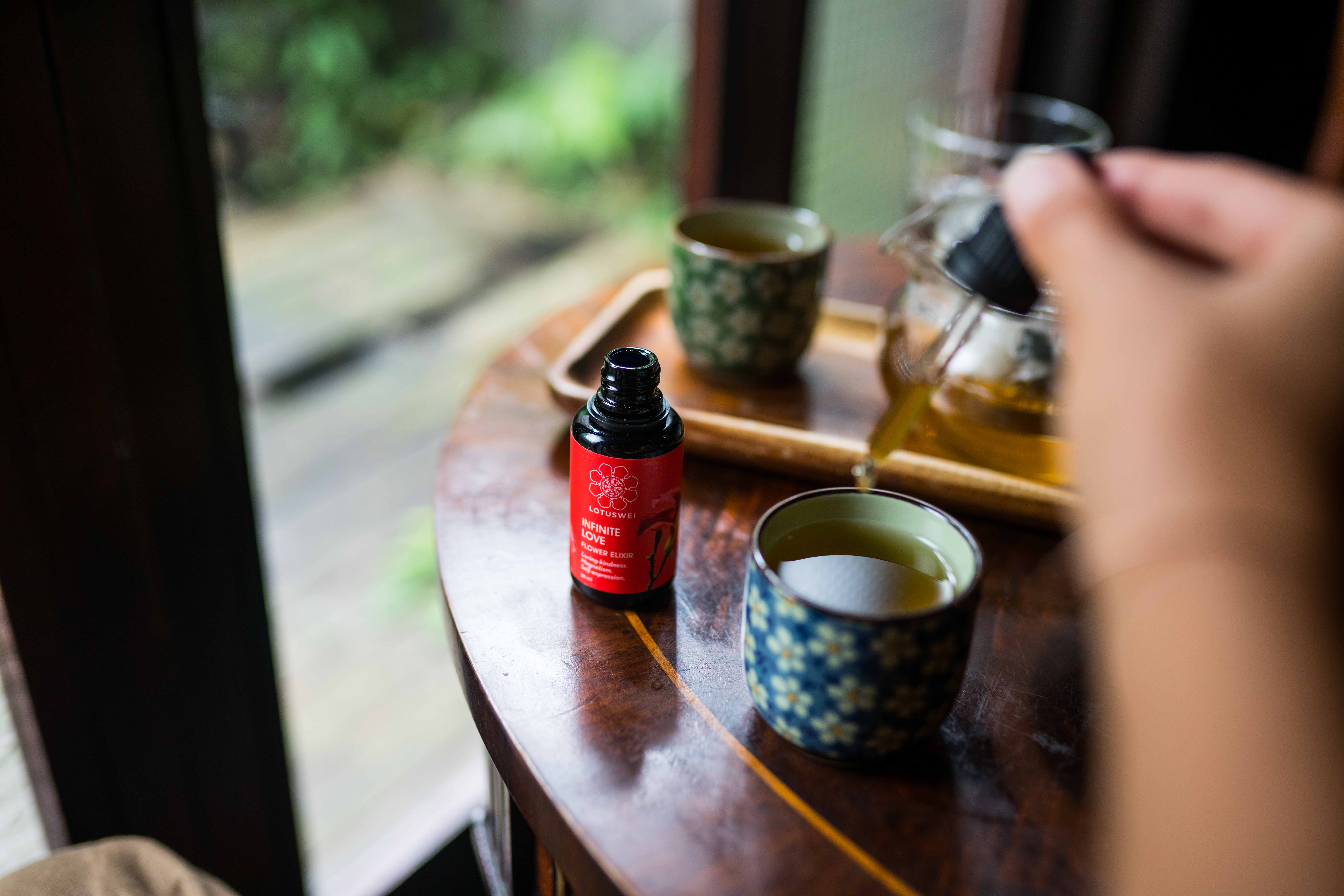 Woman adding LOTUSWEI drops to her tea sitting by scenic window view.