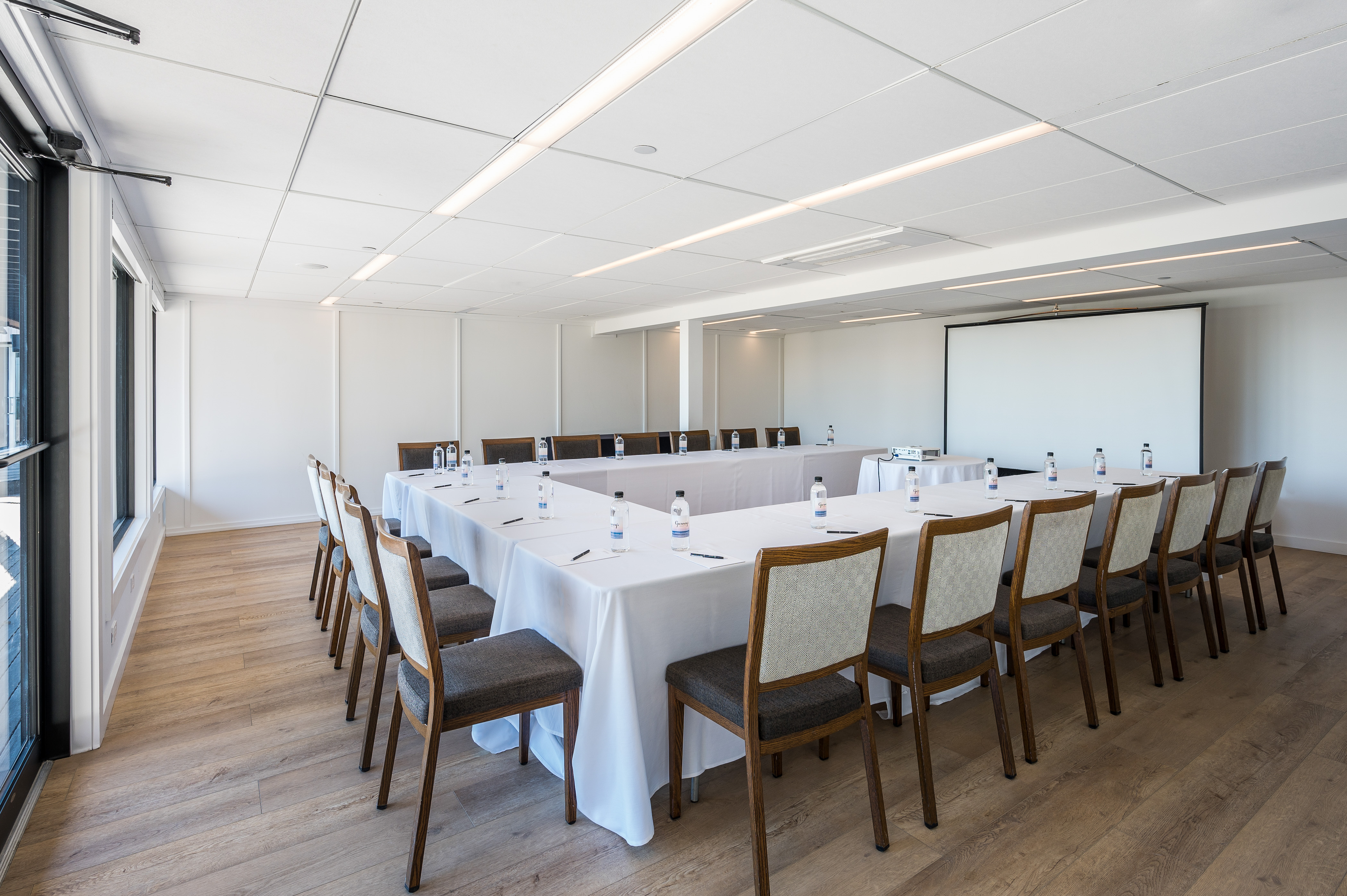 Longitude meeting venue with wood flooring, white walls, u-shaped desk setup with white linen and brown wood chairs with dark cushion. 
