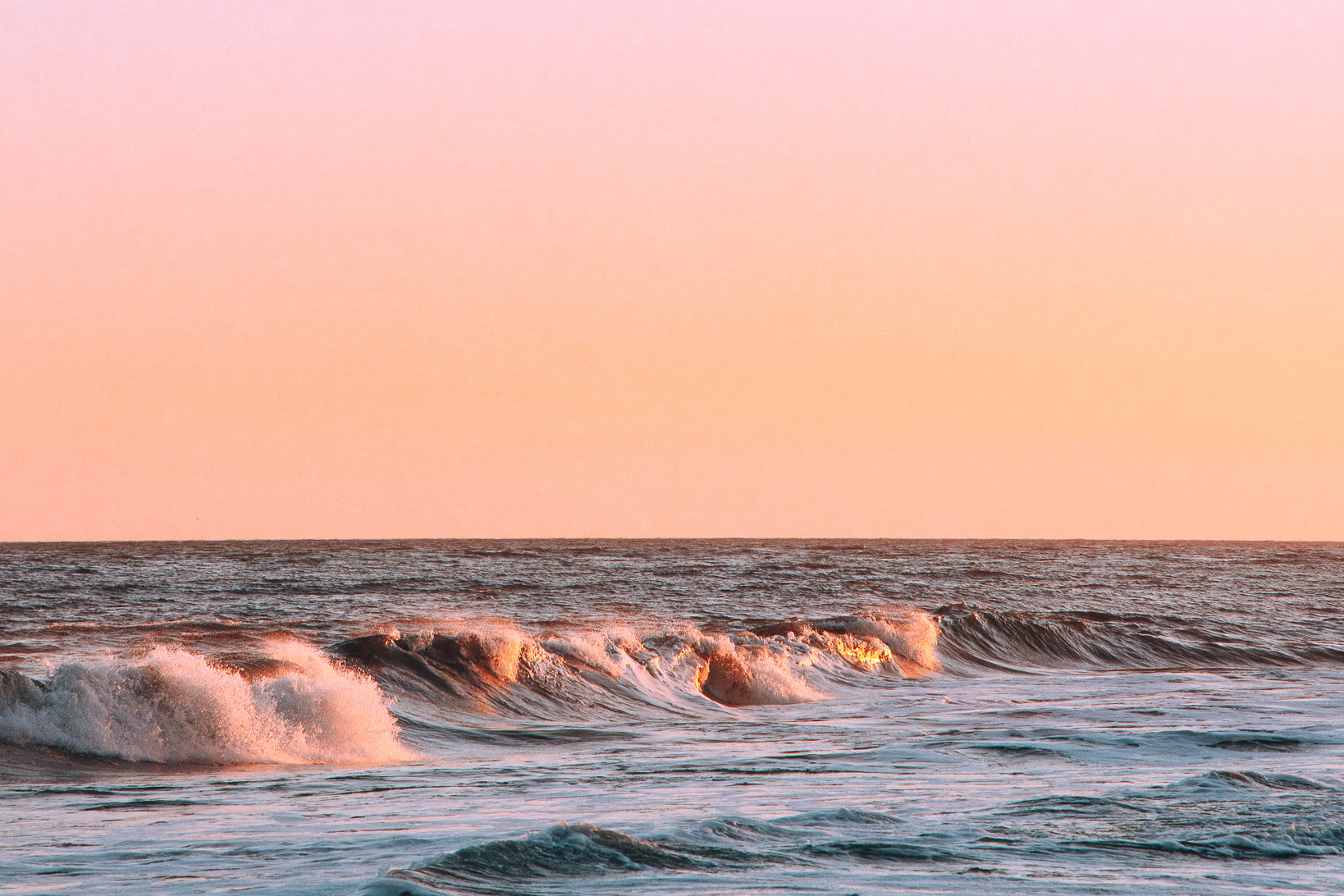 Waves crashing on shoreline during colorful, pink sunset.